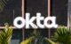 Close-up of an Okta business sign affixed to a an office building that is adorned with many palm trees