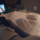 A woman lies in bed enjoying an Amazon Luna game controller as she relaxes beneath a tufted blanket.