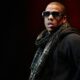 Alt Text: Jay Z wearing sunglasses, a plaid scarf, and a black hoodie ready to perform on stage at the Hove Festival.