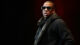 Alt Text: Jay Z wearing sunglasses, a plaid scarf, and a black hoodie ready to perform on stage at the Hove Festival.